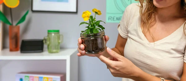 Unrecognizable curly blonde woman holding a pansy plant inside of glass pot in ecology classroom. Botanical and natural science education concept.