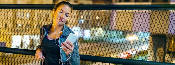 Banner of young woman runner listening music on mobile phone application on evening. Portrait of female athlete with earphones resting over bridge banister after training on town. Right copy space.