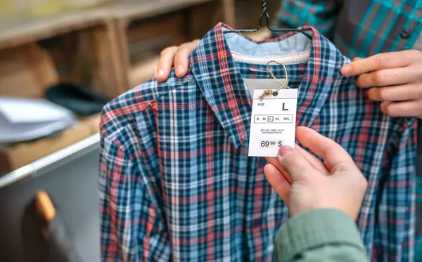 衣料品店の平らなシャツのラベルを握っている認識できない女性の顧客の手の近く 女性従業員が店の顧客にハンガーでタルタンシャツを見せています アパレルラベルに焦点を当てる — ストック写真