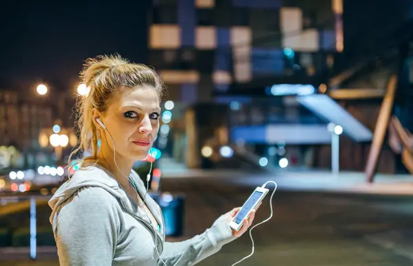Portrait of young blonde woman looking at camera while listening music on mobile phone application. Female runner with earphones holding cell with heart rate monitor display after training at night