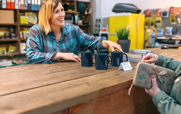 笑顔の金髪の女性従業員は ヴィンテージショップで顧客に青いマグカップを示すフリックルを持っています ローカルストアで割引カップを提供しながら 幸せなキャッシャーを購入する彼女の財布を保持している女性クライアント — ストック写真