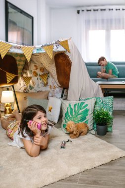 Barınak çadırında telsizle konuşan iki mutlu çocuk. Küçük kız telsizle arıyor. El yapımı çadırda, halının üzerinde kanepede oturan oğlana uzanıyor. Evde tatil kampı..