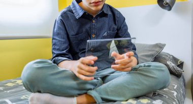 Yatağının üzerinde fütüristik tablet kullanan tanınmayan bir çocuk. Cam ekran konseptli dijital ekran. Evde taşınabilir cihaz tutan rahat genç bir çocuk..