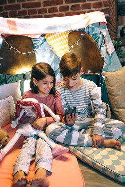 Sosyal medya başvurularında sohbet ederken cep telefonuna bakan çocuklar evdeki çadırda oturuyorlar. Mutlu küçük kız, küçük bez bebeği kucaklıyor. Yanında da rahat bir çadırda hücresini kullanıyor..