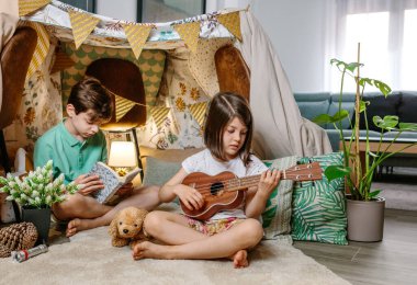 Küçük kız ukulele oynarken oğlan oturma odasında el yapımı çadır kitabı okuyor. Çocuklar evlerinde konforlu bir çadırda eğleniyorlar. Tatil kampı veya konaklama konsepti.