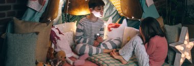 Korkmuş kızın yüzü kapalı bir şekilde evdeki karanlık sığınakta otururken çocuk korku jesti yaparken eğleniyor. Çocuklar pijama giyip ev yapımı çadırda oynuyorlar..