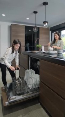 Genç bir kız bulaşık makinesinden temiz tabakları boşaltırken bir kadın modern mutfak ortamında yemek hazırlıyor, modern aletleri ve aile çalışmalarını sergiliyor.