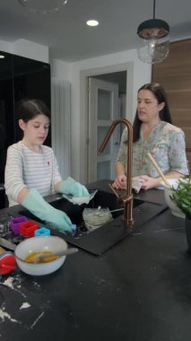 Mutfak lavabosunda bulaşık yıkayan eldiven takan mutlu bir kız. Kuruyan annesinin yanında. Modern bir mutfakta, koyu bir tezgahı ve bakır bir musluğu var. Aile paylaşımı ev işi ve kaliteli zaman.