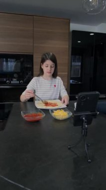 Genç bir kız pizza hamurunun üzerine domates sosu dökerken aynı zamanda modern mutfakta üç ayaklı bir telefonla yemek pişirme dersi veriyor, mutfak becerileri ve yaratıcılığını gösteriyor.