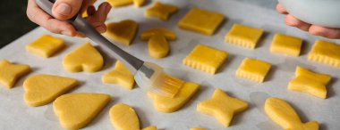 Banner of unrecognizable hands of pastry chef delicately brushing egg yolk onto raw cookies arranged on a baking sheet, skillfully preparing them for baking in the oven. clipart