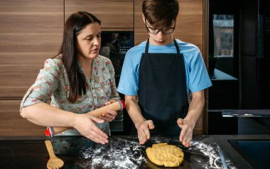 Anne ve genç oğul mutfakta kaynaşır, birlikte yemek yapmayı öğrenirler. İçindekiler ve araçlar yemek yapmak, sevgi ve iletişimi paylaşmak için etraflarını sarar. Rozetli kadın çocuğa yemek pişirmede yardım ediyor..