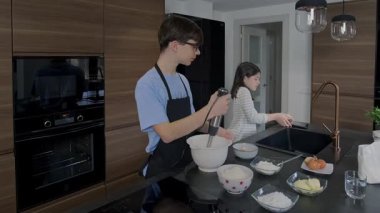 İki mutlu genç, modern bir mutfakta pasta yapıyor. Biri el çırpıcısı kullanırken diğeri de elektronik tablet tarifine başvuruyor.