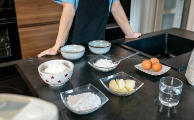 Tanımlanamayan genç pasta şefi, çağdaş bir mutfakta, parlak, koyu bir tezgahın üzerinde un, şeker, tereyağı ve yumurta kaseleri hazırlıyor.
