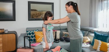 Anne ve kızının bayrağı, yaz tatili için neşe içinde kıyafet hazırlamak ve bavul hazırlamak, bikini denemek ve gülümseme paylaşmak, seyahat ve tatil maceralarının heyecanını somutlaştırmak.