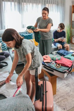 Anne ve kızı bavulları katlıyor ve paketliyor. Bu arada genç oğlu da sıcak oturma odasındaki kanepede oturup yaz tatiline hazırlanıyor. Aile yolculuk için çantaları hazırlıyor..