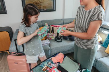 Anne ve kızı sevinçle kıyafet hazırlıyor ve yaz tatili için bavul hazırlıyor, bikinilere bakıyor ve gülümsemelerini paylaşıyor, seyahat ve tatil maceralarının heyecanını somutlaştırıyor.