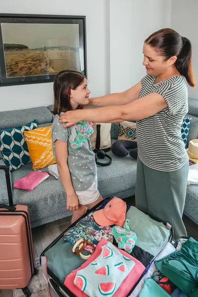 Anne ve kızı sevinçle kıyafet hazırlıyor ve yaz tatili için bavul hazırlıyor, bikinileri deniyor ve gülümsemelerini paylaşıyor, seyahat ve tatil maceralarının heyecanını somutlaştırıyor.