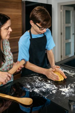 Anne ve genç oğul mutfakta kaynaşır, birlikte yemek yapmayı öğrenirler. İçindekiler ve araçlar yemek yapmak, sevgi ve iletişimi paylaşmak için etraflarını sarar. Rozetli kadın çocuğa yemek pişirmede yardım ediyor..