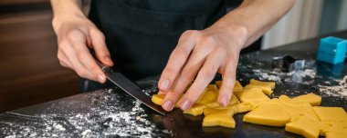 Siyah mutfak tezgahında bıçakla rulo hamurdan kalp şeklinde kurabiyeler kesen tanınmayan erkek pasta şefi pankartı.