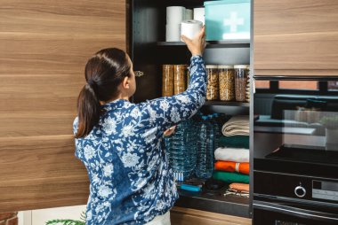 Tanınmayan bir kadın potansiyel felaketler için 72 saat boyunca hayatta kalma kiti hazırlıyor. Mutfaktaki acil durum yemek kilerine tuvalet kağıdı koyan bir kadın. Uyarı, öngörü ve kendi kendine yetme stratejisi.