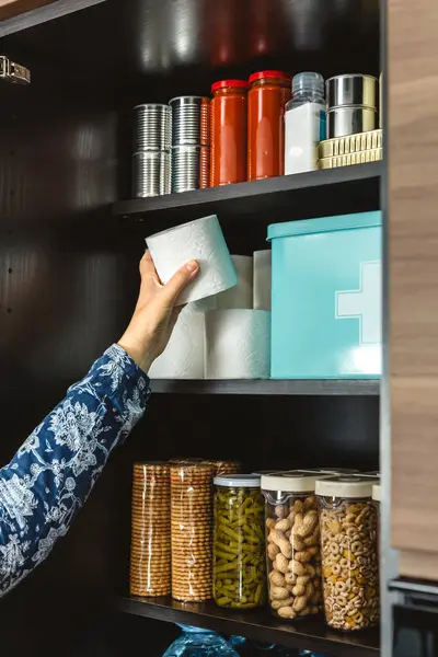 Mutfaktaki acil durum yemek kilerine tuvalet kağıdı doldurup potansiyel felaketlere karşı hazırlanan tanınmamış kadın elini kapatın. Uyanık, öngörülü ve kendi kendine yeten. 72 saat hayatta kalma kiti