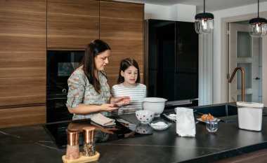 Anne ve kızı elektronik tablete bakıyor ve bir kek hazırlamak için un ve diğer malzemeleri kullanarak modern bir mutfakta tarifi takip ediyorlar. Ev kavramında aile eğlencesi.