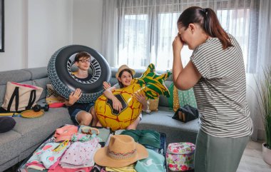 Şişme şamandıralar tutan gülümseyen çocuklar ve mutlu anneler giysi, mayo ve aksesuarlarla dolu bir valizin yanında fotoğraf çekiyorlar. Aile valizleri yaz tatili için oturma odasında hazırlanıyor..