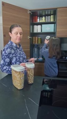 Aile, felaket durumları için mutfakta acil yemek kileri hazırlıyor. Anne ve kızı 72 saat boyunca baklagil, tahıl gevreği ve fıstık paketleri yerleştiriyor. Öngörü ve kendi kendine yetme stratejisi.