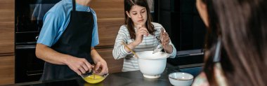 Ergen erkek ve kızın mutfakta birlikte pasta yaptığı yumurta çırpma ve pudra şekeri eklediği bir pankart. Bu arada anne tabletten yemek tarifini izliyor ve takip ediyor. Ev kavramında aile eğlencesi.