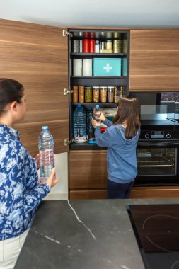 Aile, öngörü ve kendi kendine yetme stratejisinin bir parçası olarak 72 saatlik hayatta kalma kiti hazırlıyor. Mutfak kilerinde savaş ya da doğal afetler için yiyecek ve ilk yardım çantasıyla su şişelerini saklayan bir kız..