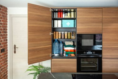 Hükümetin acil durum veya savaşa hazırlık konusundaki tavsiyelerine göre depolanan hayatta kalma kiler. Dolapta konserve yiyecekler, su şişeleri, ilk yardım malzemeleri ve modern mutfakta giysiler var..