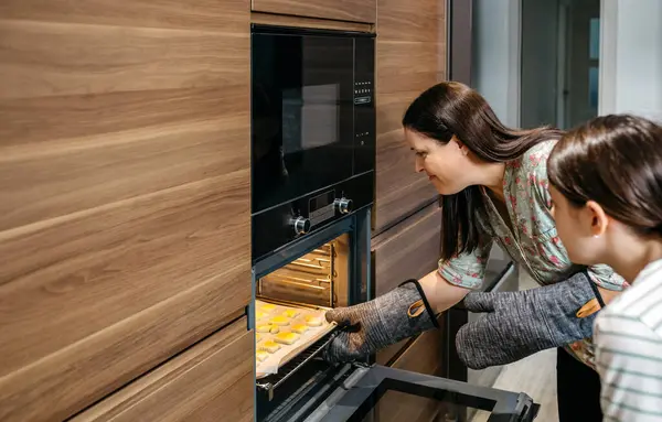 Anne ve kızı mutfaklarındaki fırından altın kurabiyelerle dolu bir tavayı çıkarıyorlar, fırın eldiveni takıyorlar ve birlikte çalışarak yeni pişirilmiş yiyecekleri dikkatlice temizliyorlar.