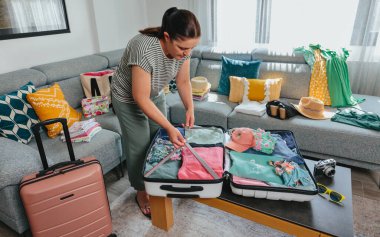 Kompresyon kayışları kullanarak valizin içinde kıyafet düzenleyen, yaz tatiline hazırlanan katlanmış giysiler ve arka plandaki koltukta yaz aksesuarları olan bir kadının portresi.