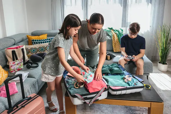 Anne ve kızı dikkatlice bavullarını masanın üstüne yerleştiriyor, genç oğlu arka plandaki dijital kamerada fotoğrafları kontrol ediyor, yaz tatiline hazırlanıyor. Yolculuk için aile çantaları..