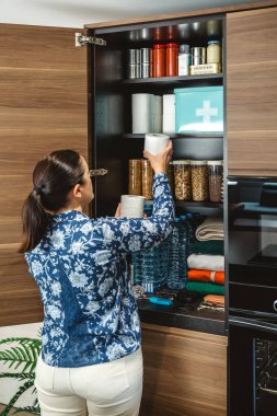 Tanınmayan bir kadın potansiyel felaketler için 72 saat boyunca hayatta kalma kiti hazırlıyor. Mutfaktaki acil durum yemek kilerine tuvalet kağıdı koyan bir kadın. Uyarı, öngörü ve kendi kendine yetme stratejisi.
