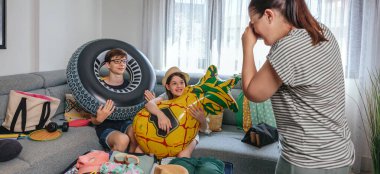 Şişme şamandıralar tutan gülümseyen çocuklar ve mutlu anneler giysi, mayo ve aksesuarlarla dolu bir valizin yanında fotoğraf çekiyorlar. Aile valizleri yaz tatili için oturma odasında hazırlanıyor..