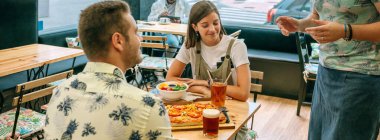 Pizza, salata ve içeceklerle masada otururken restoranda garsona yemek ısmarlayan mutlu çiftin bayrağı ve arka planda oturan başka bir müşteri..