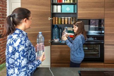 Anne ve kızı mutfak kilerinde su şişeleri, yiyecek ve erzak depoluyorlar. Aile 72 saatlik savaş ve doğal afetler için hayatta kalma kiti hazırlıyor. Uyarı, öngörü ve kendi kendine yetme stratejisi.