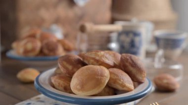  Altın Hollanda Poffertjes şuruplu ve şekerli. Videoda, pudra şekeri beyaz bir tabakta mavi desenli mini keklerin üzerine dökülüyor.