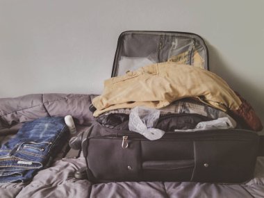 Yatağın üstünde siyah bir bavul ve giysiler olan küçük bir oda.