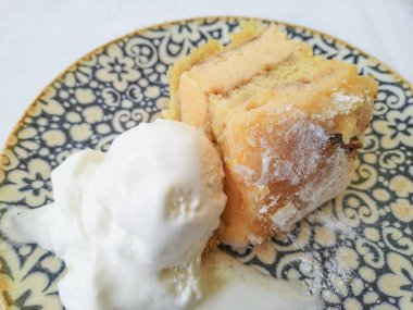 Yüksek açılı, toz şekerle tozlanmış iki parça sünger keki mavi ve beyaz dekoratif seramik tabakta bir kepçe vanilyalı dondurmayla servis ediliyor. Doğal ışıklandırma ve rahat, kırsal bir atmosfer..