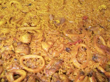 Kalamar, pirinç ve et parçalarıyla geleneksel İspanyol paellasını yakından çek. Doğal ışık, altın tonları ve dokuları ortaya çıkarır. Renk ve lezzet dolu klasik bir Akdeniz yemeği..
