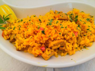 Yüksek açılı safran sarısı pirinç, tavuk parçaları ve sebzelerle birlikte, beyaz oval bir tabakta servis ediliyor. Canlı renkler ve süslü otlar sıcaklığı, besinleri ve ev yapımı lezzeti çağrıştırıyor.