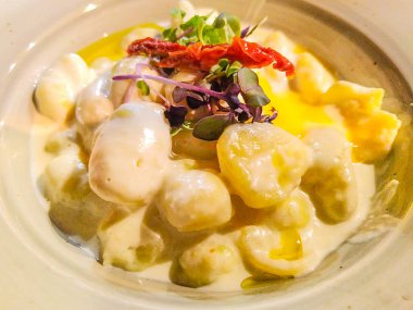 Kurutulmuş domates ve taze mikroyeşilliklerle süslenmiş kremalı sosla kaplı bir kase gnocchi 'nin yüksek açılı görüntüsü. Zarif kaplama ve sıcak ışıklandırma rafine bir konfor ve gurme ev yemeği hissi yaratır.