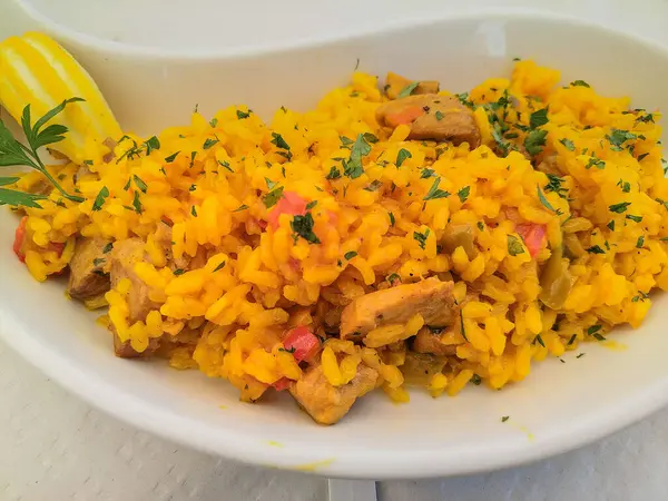 Yüksek açılı safran sarısı pirinç, tavuk parçaları ve sebzelerle birlikte, beyaz oval bir tabakta servis ediliyor. Canlı renkler ve süslü otlar sıcaklığı, besinleri ve ev yapımı lezzeti çağrıştırıyor.