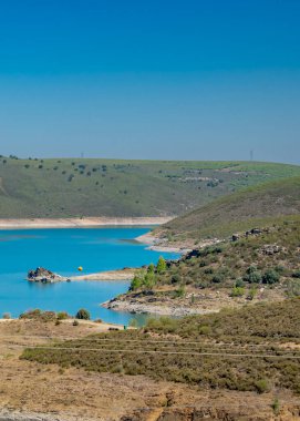 Yuvarlanan tepelerin arasında kurumuş arazi ve en az bitki örtüsü olan sakin mavi gölün göz hizasında görüntüsü..
