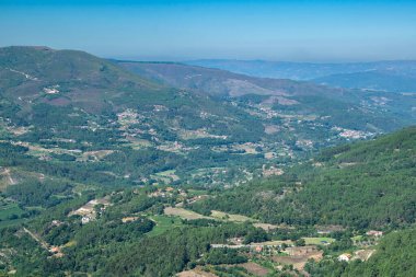 Yoğun ormanları, dağınık evleri ve bulanık mavi gökyüzünün altında dolambaçlı yolları olan engin bir dağ vadisinin yüksek açılı manzarası.