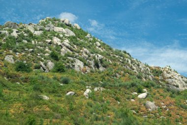 A shot of a rocky hilltop with sparse green vegetation against a blue sky with wispy clouds. 