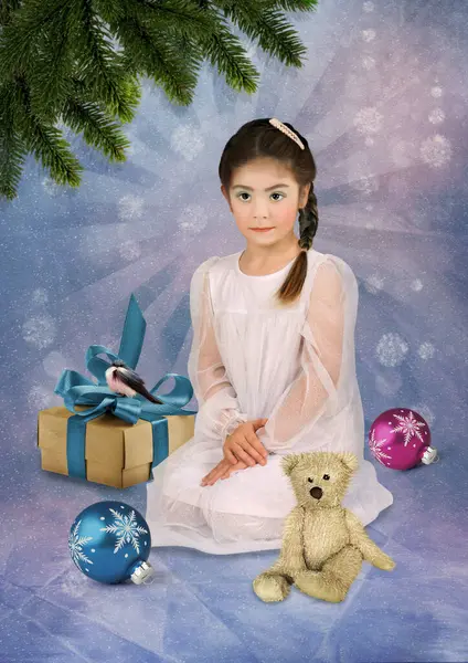 穿着白色连衣裙的小女孩坐在她的腿上 一只泰迪熊在冬季的背景下被圣诞礼物环绕着 图库图片