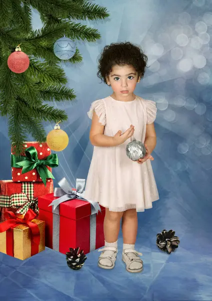 在圣诞树下 一个手里拿着圣诞球的小女孩被圣诞树下的礼物环绕着 图库图片